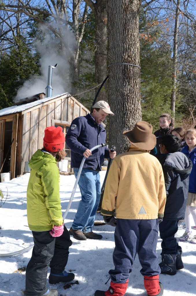 A man demonstrates maple syrup production to a group of children in a snowy outdoor setting, with steam rising from a nearby sugar shack. The children are dressed warmly for winter, showing interest in the process.