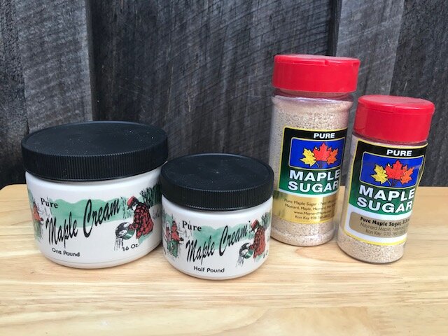 Two jars of pure maple cream, one labeled as one pound and the other as half pound, alongside two containers of pure maple sugar, set against a rustic wooden background. Ideal for cooking and baking enthusiasts.